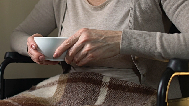 Mature woman sitting near window in wheelchair drinking hot tea, home coziness